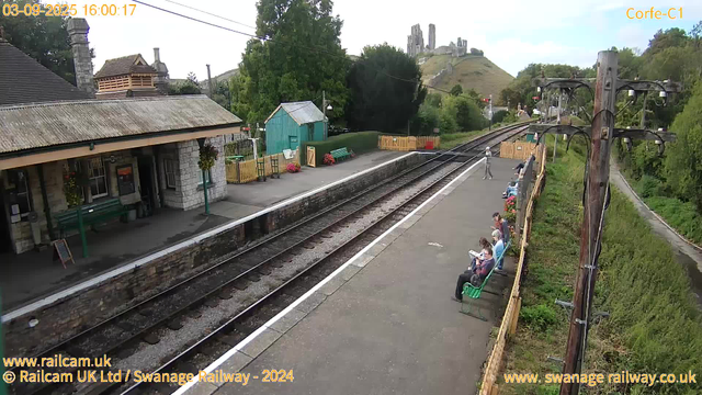 A small train station platform with a stone building on the left, decorated with hanging flower baskets. Several people are seated on green benches along the right side of the platform, some reading or using their phones. A single person stands near the center of the platform, and others are sitting further down. There is a wooden fence with flower pots and a small green shed in the background. Railway tracks run parallel to the platform, disappearing into the distance with lush greenery on both sides. In the far background, a hill with a ruined castle or similar historic site is visible on top. A large wooden utility pole with multiple wires stands on the right edge of the image. The sky is partly cloudy with some blue showing through. The timestamp shows the date as September 3, 2025, at 16:00:17.