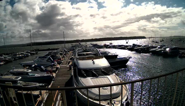 A marina filled with various boats and yachts docked along wooden piers. The water is calm with sunlight reflecting off its surface, and there is a cloudy sky overhead. A black railing is visible in the foreground. The horizon shows more boats and a distant shoreline.