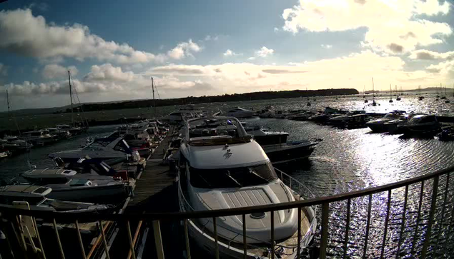A marina filled with numerous yachts and boats moored along wooden docks, with a view of the open water reflecting sunlight, under a partly cloudy sky with the sun low on the horizon. A railing is visible in the foreground.