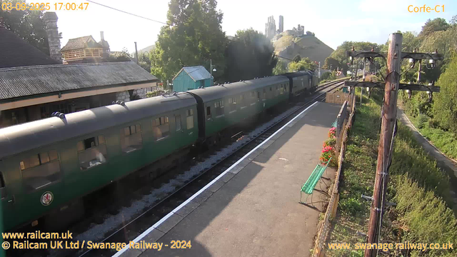 A green train with several carriages is stopped at a small railway station platform. The platform has a green bench and a row of potted pink flowers along the railing. There are wooden utility poles with electrical wires running overhead. In the background, there are trees, a blue building, houses with tiled roofs, and a hill with a castle or ruins on top. The timestamp on the image reads "03-09-2025 17:00:47". The image is from the Swanage Railway, with website links at the bottom.