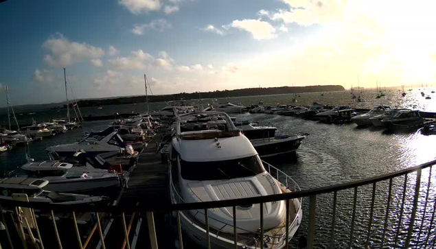 A marina filled with various boats and yachts docked along the pier, with the sun setting in the distance, casting a warm glow over the water. The water reflects the sky and boats, and there is a curved railing in the foreground.
