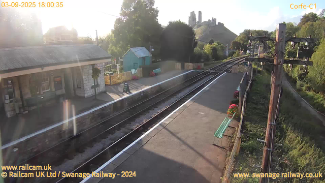 A small train station platform with a building on the left, decorated with hanging flowers and a bench. The railway tracks run through the center, extending towards the background. To the right, there are wooden utility poles with electrical wires and flower pots attached to a railing. Behind the platform is a lush, green landscape with trees and a hill featuring a castle ruin at the top. The sky is mostly clear with a few clouds, and the sunlight is casting long shadows.