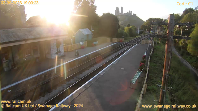 A railway station platform bathed in early evening sunlight with the sun low in the sky, causing lens flare. There are train tracks running parallel to the platform, which has colorful flowers in pots along the edge. A small building with a pitched roof is situated next to the platform, and a fenced-off area is visible behind it. On the right, wooden utility poles with multiple electrical wires line the platform. In the background, a hill with castle ruins or old stone structures is visible atop a green hill. The timestamp indicates the date and time as September 3, 2025, at 19:00:23.