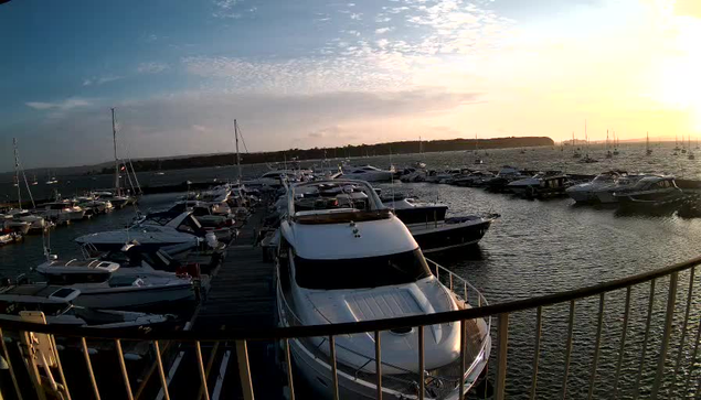 A marina filled with numerous boats and yachts docked along the water, with the sky showing a sunset or sunrise casting a warm glow and clouds scattered across the sky. The image is taken from a balcony or platform with a curved railing in the foreground.