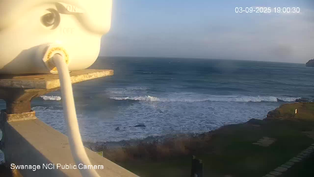 A view of the ocean with waves breaking near the shore, seen from a high vantage point on a coastal area. In the foreground, part of a white device or camera installation is visible, and the sky is mostly clear with some clouds. The timestamp on the image reads "03-09-2025 19:00:30," and the bottom left corner indicates it is from the "Swanage NCI Public Camera."