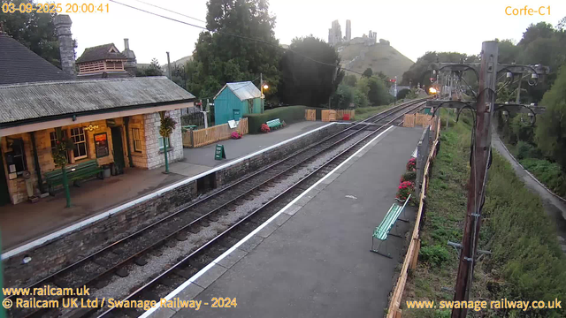 A small train station platform with a building on the left, decorated with hanging flower baskets and green benches. There are some flower pots along the edge of the platform, and a green "Way Out" sign near the building. Overhead, electrical lines run across the scene. To the right, a utility pole supports electrical wires, and a narrow path runs alongside the platform with more flower pots and a green bench. In the background, a hill with ruins or a castle is visible, and trees surround the area. The sky appears to be overcast or approaching sunset. The image has a timestamp of September 3, 2025, at 8:00 PM.