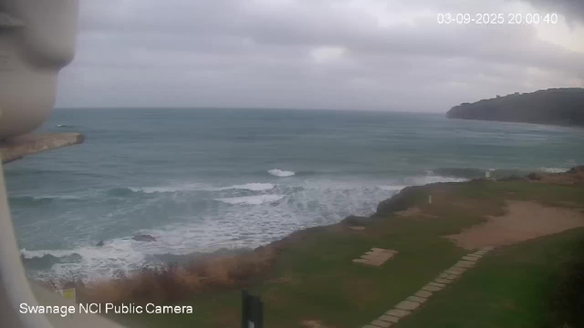 A view of the ocean with waves breaking on the shoreline, under a cloudy sky. There is a grassy area and a stone pathway on the right side, with a distant landmass or hill in the background. The image appears to be taken from a webcam at dusk or evening.