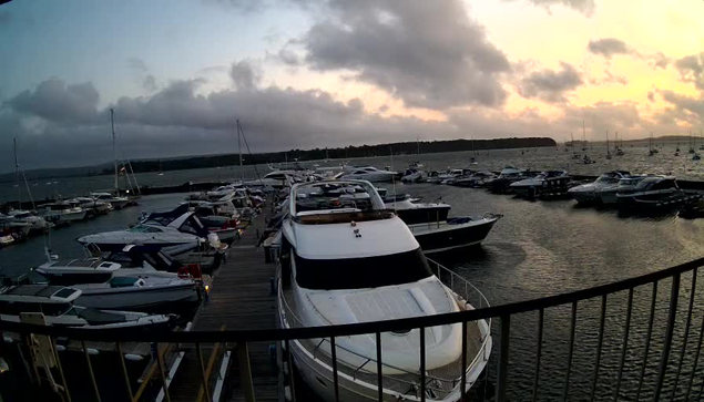 A marina with numerous boats and yachts docked along wooden piers. The water is calm, and the sky is partly cloudy with the sun setting, casting a warm glow on the scene. There is a black metal railing in the foreground.