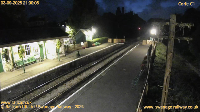 A nighttime view of a small train station platform with a building illuminated on the left side. The platform has benches, potted plants, and a small shelter. There are railway tracks running through the center of the image, and a pathway with a wooden fence on the right side. The sky is dark with some clouds visible. The photo includes the date and time in the top left corner, and the website and company information are displayed at the bottom.