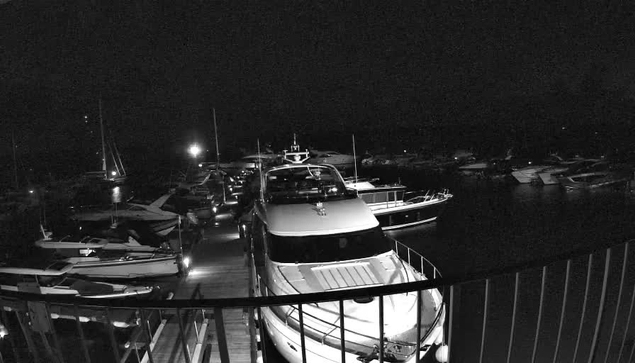 A nighttime scene at a marina shows multiple boats docked along the water. In the foreground, a large yacht is visible with a railing in front. Several smaller boats are lined up to the left and right, with their masts and hulls partly illuminated by nearby lights. In the background, more boats are docked, and the dark sky is barely visible against the still water. A bright light source is near the center, casting reflections on the water's surface.