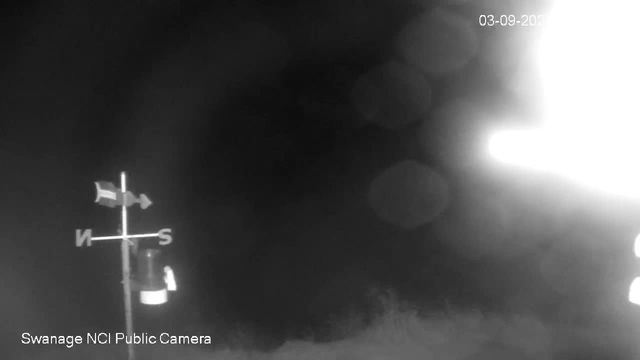 A black-and-white image from a webcam shows a dark outdoor scene with a weather vanes and a small object near it, possibly a container or light fixture, on the left side. The image has large bright glare or overexposure on the right side. The top right corner displays the date "03-09-20," and the bottom left corner reads "Swanage NCI Public Camera."