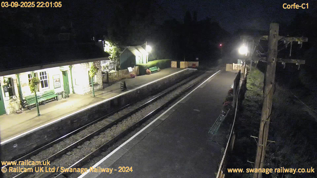 A quiet railway station platform at night, illuminated by a few bright lights. The station building on the left has white walls and green doors, with hanging flower baskets beneath the windows. A green bench and some flower arrangements are present on the platform. There are wooden barriers at the far end of the platform. On the right, an electric pole with multiple wires runs alongside the track, which is empty and stretches into the dark. The timestamp in the top left corner shows the date and time as "03-09-2025 22:01:05."