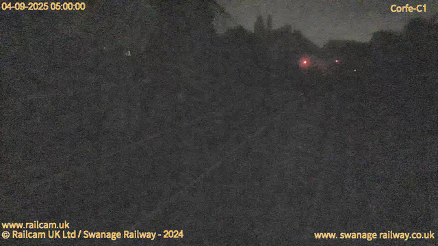 A dark, nighttime landscape with very limited visibility. In the distance, there are a few faint red lights, possibly from distant structures or signals. The image appears grainy, indicating low light conditions. Text overlays indicate the date and time as April 9, 2025, 05:00:00, and mention the location as Corfe-C1, with references to Railcam UK Ltd and Swanage Railway.