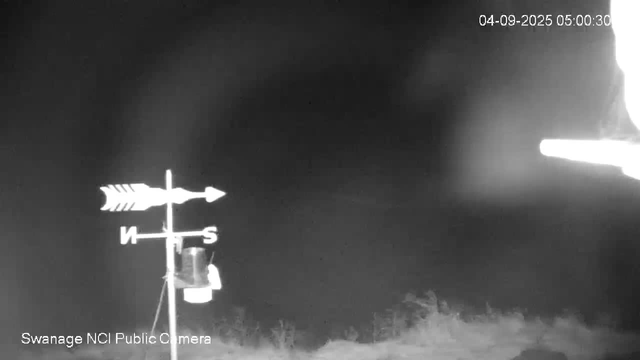 A nighttime black-and-white image showing an outdoor scene with a weather vane in the foreground. The weather vane has an arrow pointing east and west, with the letters N, S, E, W indicating the directions. There appears to be some equipment or objects attached to the weather vane stand. The sky is dark with no visible stars or moon, and the scene is faintly illuminated. The timestamp in the upper right corner reads "04-09-2025 05:00:30."