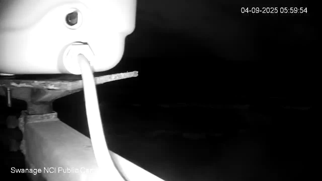 A close-up image from a webcam shows a white device, possibly a sensor or camera, mounted on a metal platform. The background is dark, and a date and time stamp reads "04-09-2025 05:59:54" in the upper right corner. The lower left corner has the text "Swanage NCI Public".