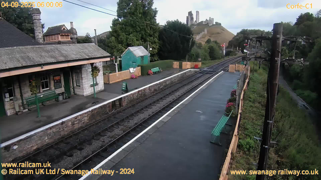 A small train station platform with a stone building on the left that has green benches, hanging flower baskets, and a notice board. There are two green benches along the platform, and a green shed with pink flowers nearby. The railway tracks run through the center, with a safety out sign near the shed. Orange and yellow safety barriers are at the far end of the platform. To the right, there's a green fence with flower pots, and electricity poles with overhead cables run parallel to the tracks. In the background, a hill with ancient castle ruins sits atop. The sky is cloudy, and it is early morning, as indicated by the timestamp.