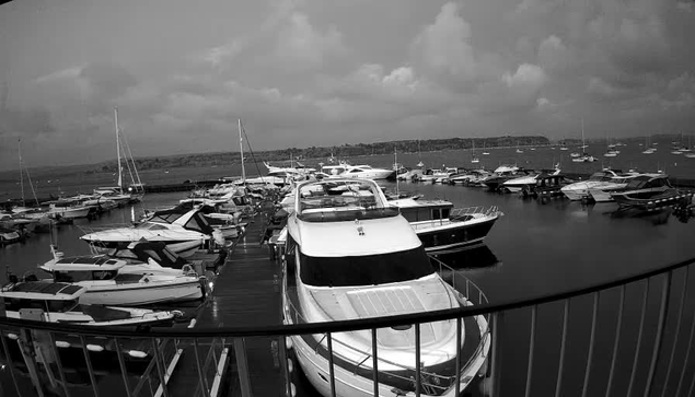 A black and white photo of a marina filled with numerous boats and yachts docked alongside a pier. The scene includes several sailboats and motorboats on the water, with a cloudy sky overhead and distant land in the background. A curved safety railing is visible in the foreground.