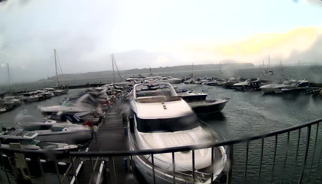A view from a webcam showing a marina with numerous boats and yachts docked at the piers, with calm water and a cloudy sky in the background. Some boats are large and modern, while others are smaller. The scene appears to be during a rainy or misty day, as there are streaks of water on the camera lens and the overall lighting is subdued.