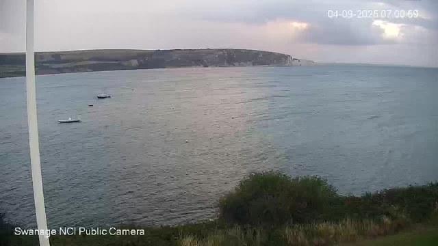 A wide view of a coastal scene showing a calm body of water with three small boats floating near the shoreline. In the background, there are white chalk cliffs and a cloudy sky with patches of sunlight breaking through. The image has a timestamp in the upper right corner and a label in the lower left corner reading "Swanage NCI Public Camera."