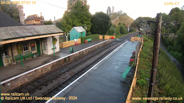 A small railway station platform with a stone building on the left, decorated with hanging flower baskets and green benches. There is a green shed near the center, surrounded by a yellow picket fence with pink flowers. The platform is empty and appears to be wet, possibly from recent rain. Overhead power lines run along the right side, supported by wooden poles. In the background, there are lush green trees and a hillside with a castle or ruins at the top. The time stamp shows early morning, and the sky is light.