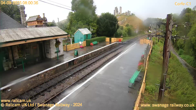 A rain-soaked railway platform with a building on the left featuring a chimney and hanging flower baskets. The platform has a green bench and a "Way Out" sign, with colorful flower pots near the fence. Railway tracks extend into the distance, with oncoming rain creating a misty appearance. A tall utility pole with multiple wires is on the right, and lush green trees line the right side of the scene. In the background, a hilltop castle or ruins is visible through the mist. The timestamp indicates the scene was captured at 8:00 am on September 4, 2025.