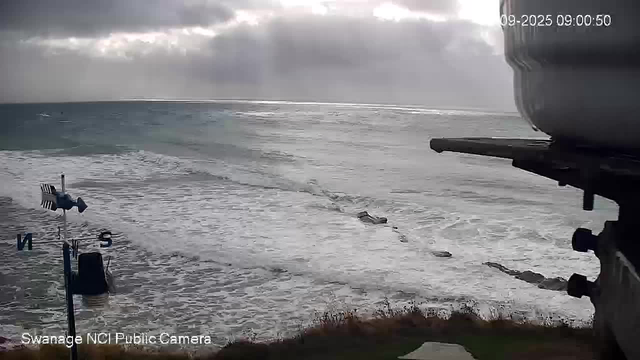 A view of the ocean with waves crashing onto the shore under a cloudy sky. A boat is in the water, moving away from the shore, leaving a wake behind it. There is a weather vane with directional indicators (N, S, E, W) visible in the foreground on the left side. Part of a large structure or equipment is visible on the right side of the image. The timestamp in the top right corner shows "2025-09-09 09:00:50."