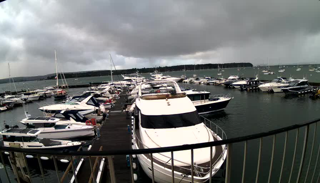 A marina filled with numerous docked boats and yachts under a cloudy, dark sky. A wooden pier extends into the water, with a large white yacht prominently in the foreground. The water is calm, and the horizon shows a distant landmass with a long, dark shoreline. The overall atmosphere appears overcast and slightly stormy.