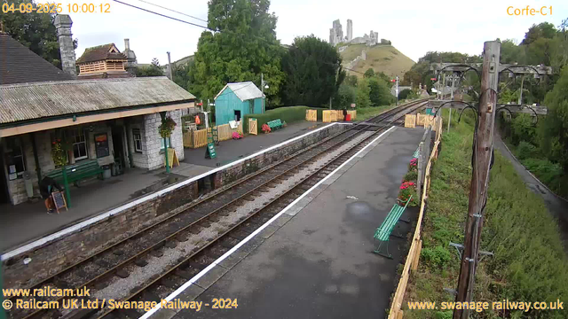 A small railway station platform with a building on the left, decorated with potted plants and hanging baskets. There are green benches and a small blue shed near the edge of the platform. A woman is reading a menu or signboard near the building. The railway tracks run parallel to the platform, extending into the distance. To the right, a wooden fence with flower pots separates the platform from a grassy area with trees. An old wooden utility pole with electrical wires is situated on the right side. In the background, a hill features a castle or ruins atop it, and a castle tower is visible in the distance. The timestamp indicates the photo was taken on September 4, 2025, at 10:00 AM.