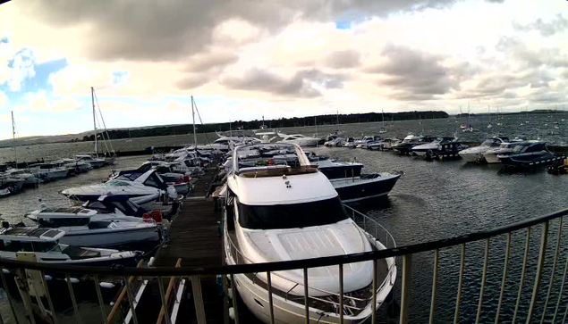 A view of a marina with multiple boats and yachts docked along wooden piers. The water is slightly choppy, and the sky is overcast with dark clouds. The scene is captured from a vantage point behind a curved railing.