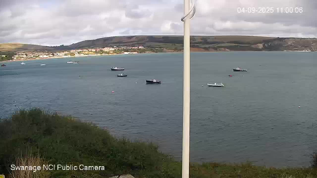 A view of a calm bay with several boats anchored in the water. In the background, there are hills with a small town or village along the shoreline. The sky is partly cloudy, and the timestamp shows "04-09-2025 11:00:06." The bottom left corner has the text "Swanage NCI Public Camera."