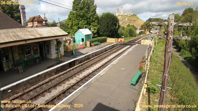 A small train station platform with a building on the left that has a stone facade and a corrugated metal roof. There are flower arrangements hanging on the building and a chalkboard sign on display. In the middle background, there are colorful flower beds, a green bench, and a small turquoise shed. Beyond the shed, a lush green hedge and large trees are visible. To the right, a wooden utility pole with electrical wires runs along a narrow, tree-lined pathway. In the distance, a hill with a castle ruin on top rises above the landscape. The sky is partly cloudy with patches of blue, and the scene is brightly lit, indicating daytime. The top leave indicates the date and time: September 4, 2025, at 11:00:12.
