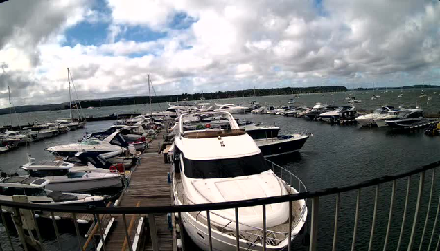 A marina filled with numerous boats and yachts docked along wooden piers. The water is calm, and the sky above is partly cloudy with patches of blue visible. In the foreground, a large white yacht is docked, with a metal railing partially obstructing the view.