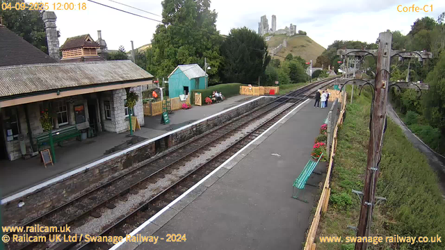 A small railway station platform with a building on the left, decorated with hanging flower baskets. There are some benches, a green trash bin, and a green sign indicating the exit. A few visitors are sitting on benches or standing near the platform, which is bordered by a yellow fence. A couple of people are standing together further down the platform. To the right, there are power lines and a wooden utility pole. In the background, a hill with ruins of an old castle or fortress is visible, surrounded by trees and greenery. The scene is captured during daytime on April 9, 2025.