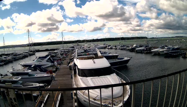 A marina with numerous boats and yachts docked along wooden piers, set against a backdrop of a cloudy sky and distant land on the horizon. The image is captured from a viewpoint behind a curved railing.